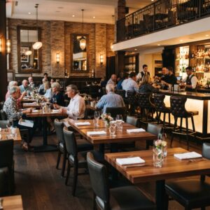 Modern upscale restaurant dining area with warm lighting, dark wood tables, and blue velvet chairs, featuring a bustling bar in the background.